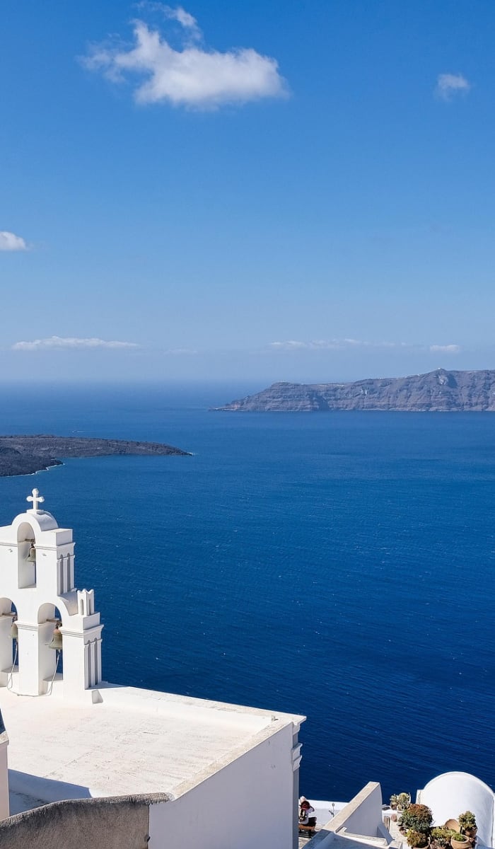 White buildings with a blue dome and bell tower overlooking the deep blue sea and distant islands under a clear sky in Santorini, Greece.