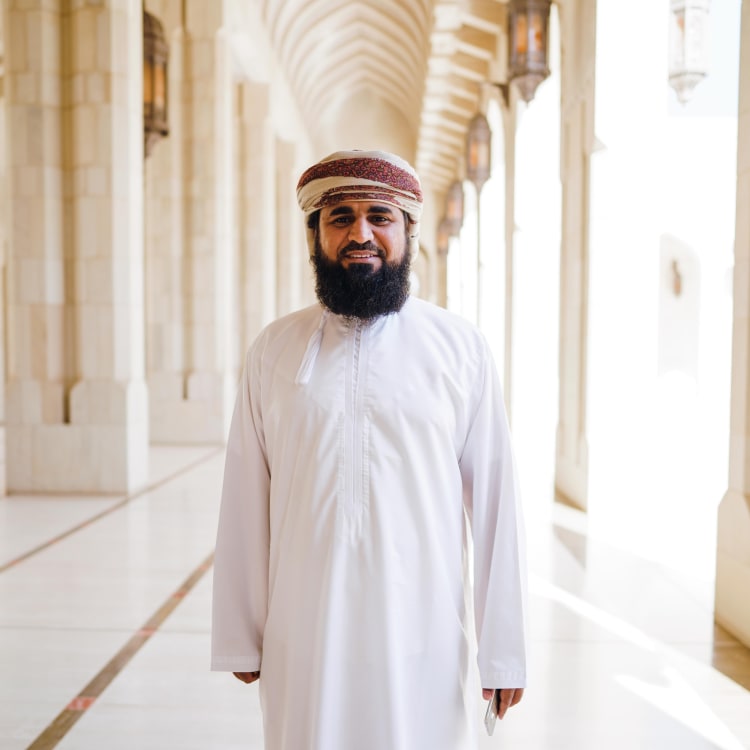 A man in traditional Omani attire standing inside a grand mosque with arched corridors and hanging lanterns.