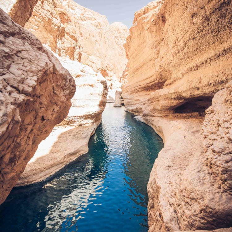 Ein enger blauer Wasserweg, der sich durch steile, sandige Canyonwände in Wadi Shab, Oman, schlängelt.