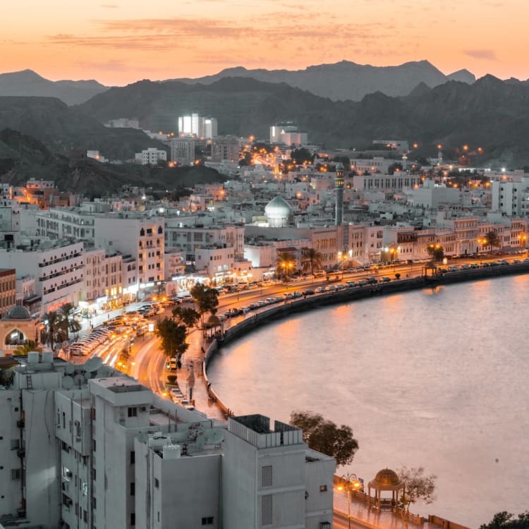 Dusk over Muscat's waterfront with illuminated buildings and mountainous backdrop.