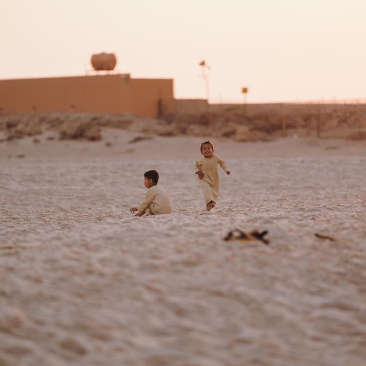 Two children playing in the sand on a desert landscape in Oman with a building and water tank in the background.