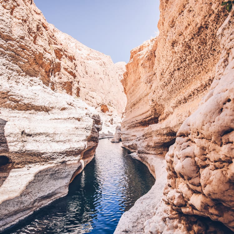 Ein enger Wasserweg mit klarem blauem Wasser, das zwischen hohen, geschichteten beige- und braunfarbenen Felsformationen unter einem hellen Himmel fließt.