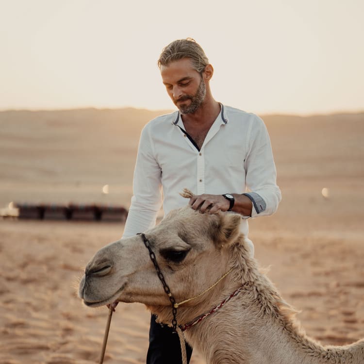 A man in a white shirt with rolled-up sleeves stands in a desert, holding a camel's rope, with the camel in the foreground and desert landscape in the background.
