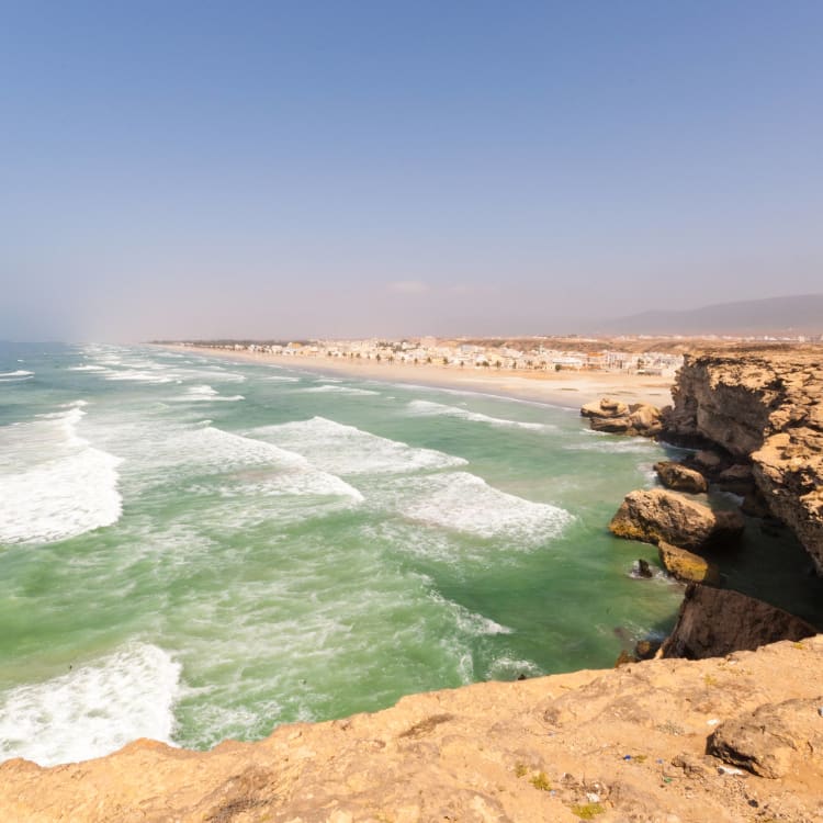 Ein Küstenblick auf den Salalah-Strand in Oman mit Wellen, die gegen felsige Klippen schlagen, unter einem klaren blauen Himmel.
