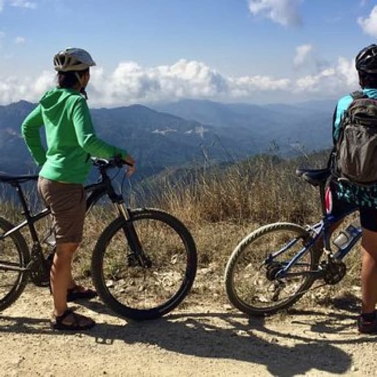 Zwei Mountainbiker stehen auf einem Schotterweg mit Blick auf eine malerische Berglandschaft und teilweise bewölkten Himmel am Tag.
