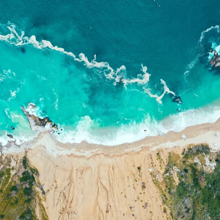 Luftaufnahme eines Sandstrands, der auf lebhafte türkisfarbene Ozeanwellen trifft, mit Felsen und Grün am Rand.