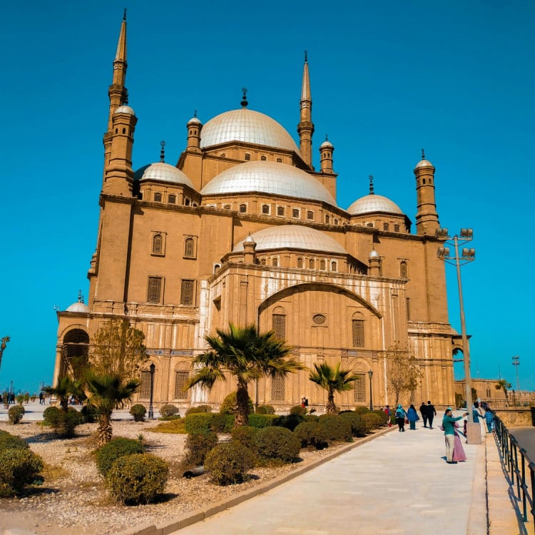 Exterior view of the historic Mosque of Muhammad Ali in Cairo under a clear blue sky, with visitors walking along the pathway and landscaped greenery in the foreground.