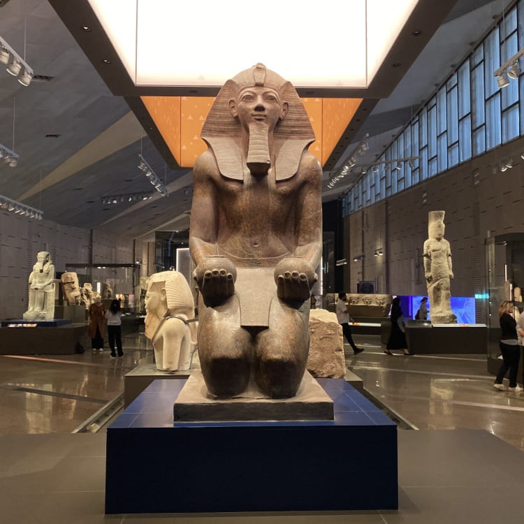 Eine große Steinstatue eines altägyptischen Pharaos, der auf einem Thron sitzt, ausgestellt in einer geräumigen Museumshalle mit anderen ägyptischen Artefakten und mehreren Besuchern.