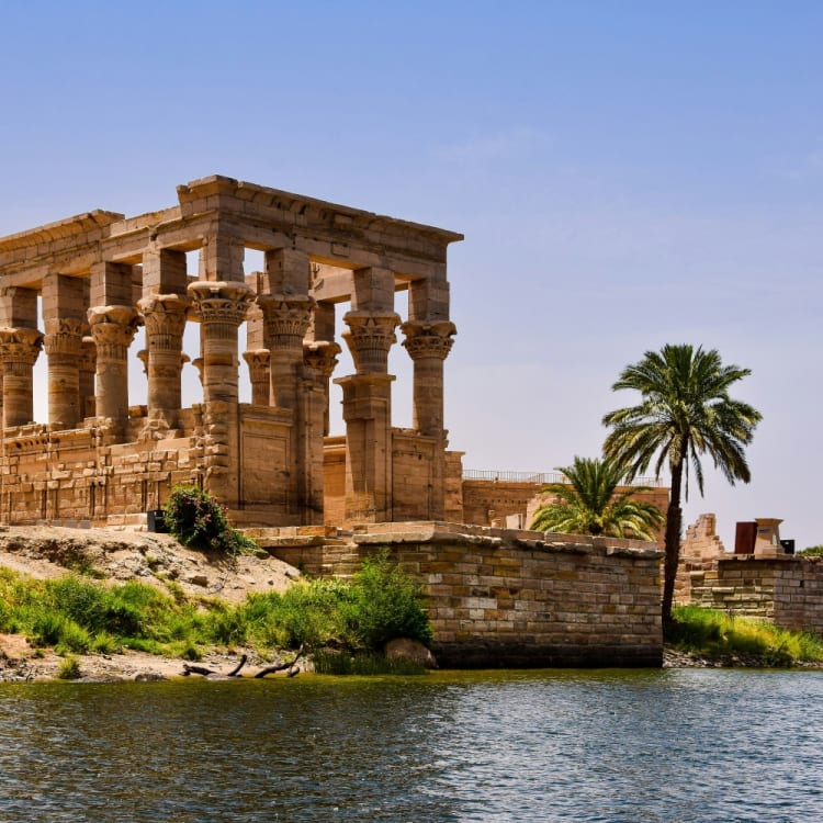 Ancient Egyptian temple with tall columns located by the riverbank, with palm trees nearby.