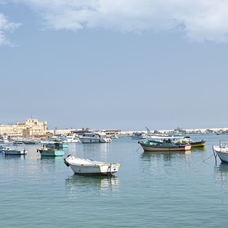 Mehrere kleine Boote und Yachten liegen in ruhigem Wasser im Hafen von Bahary, Alexandria, mit der Zitadelle von Qaitbay im Hintergrund unter einem teils bewölkten Himmel.