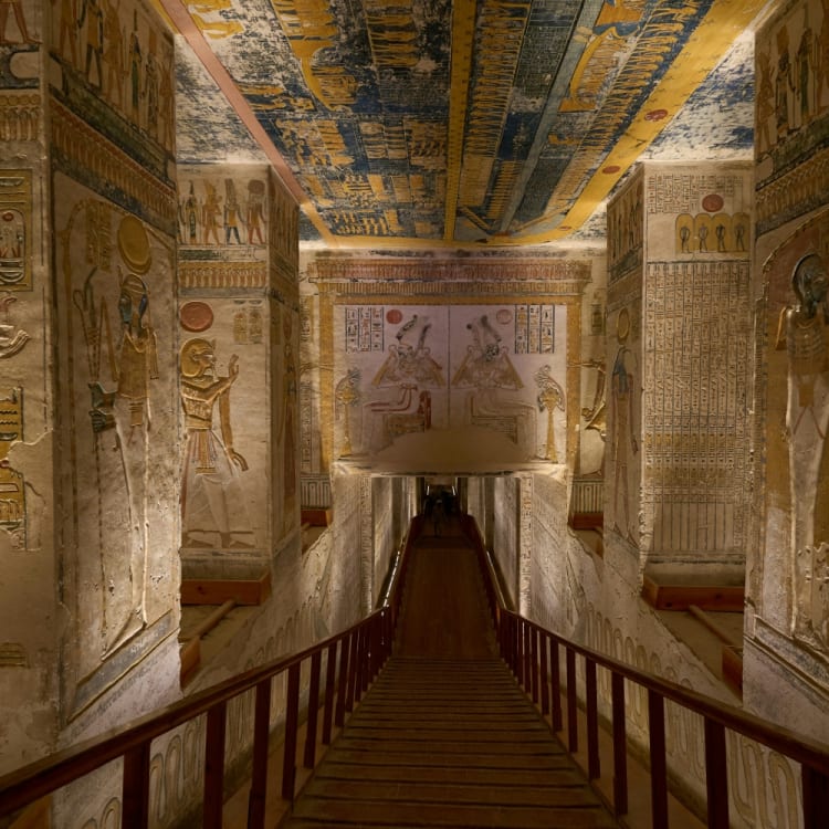 A narrow wooden walkway inside the tombs of the Valley of the Kings in Luxor, Egypt, lined with ancient Egyptian wall carvings and hieroglyphs.