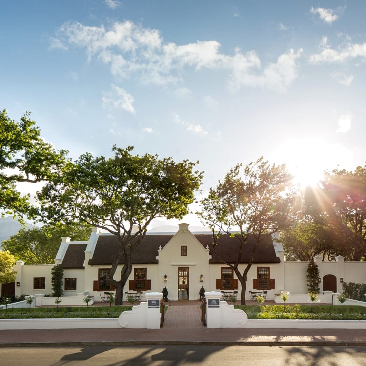 A charming white hotel with a dark roof, surrounded by lush trees and a well-maintained garden, sunlight illuminating the scene from the right.