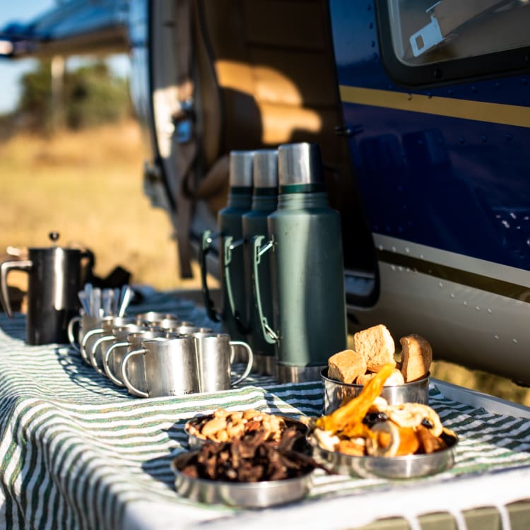 Ein Tisch, der mit einem grün-weiß gestreiften Tuch bedeckt ist, steht neben einem Hubschrauber in einer offenen Graslandschaft. Auf dem Tisch sind mehrere Metalltassen, Thermoskannen, eine Kaffeekanne und Schüsseln mit verschiedenen Snacks wie getrockneten Früchten und Brotstücken arrangiert.