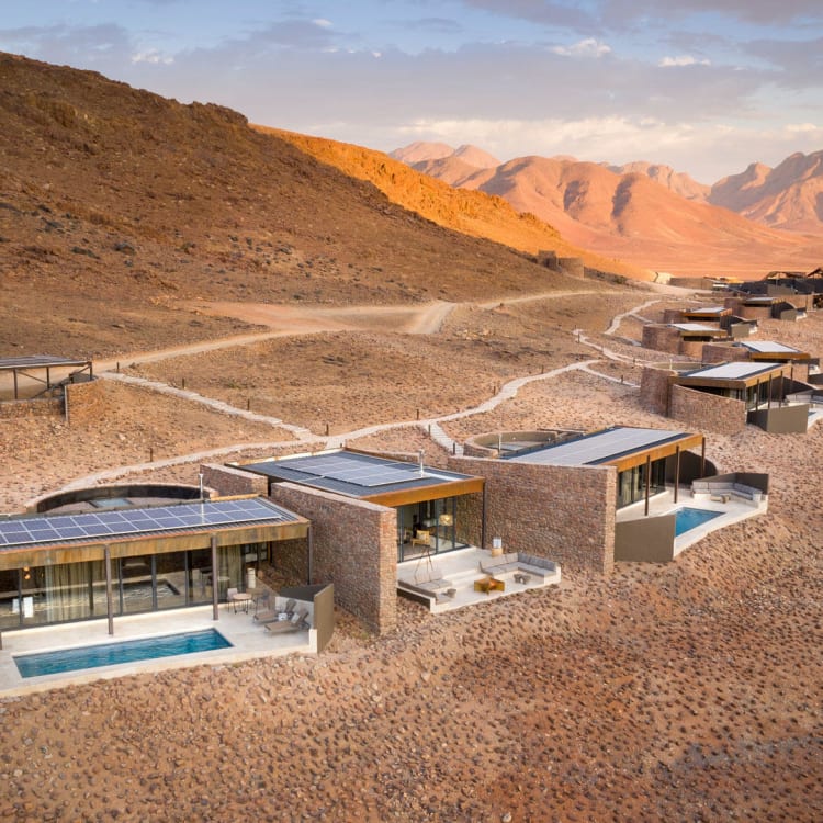 A series of modern desert lodges with swimming pools extend across the arid landscape of Namibia's Namib Desert, with mountains in the background.