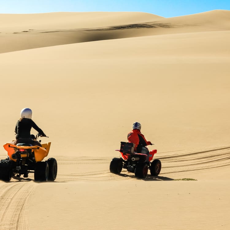 Zwei Personen fahren Quad durch die sandigen Dünen einer Wüste in Namibia.