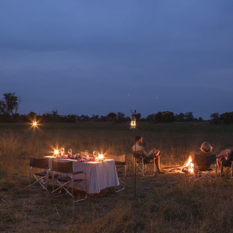 In der Dämmerung, unter einem tiefblauen Himmel, sitzen drei Personen um ein Lagerfeuer auf Campingstühlen in einer weitläufigen Graslandschaft. Vor ihnen stehen zwei Esstische, die festlich mit weißen Tischdecken und Gläsern gedeckt sind.