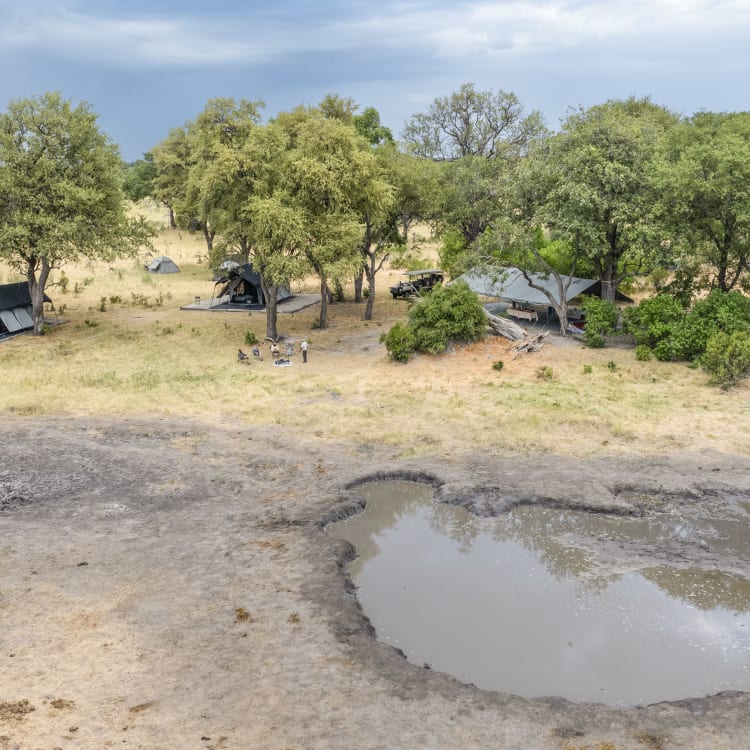 Eine Luftaufnahme eines Safari-Camps, das in einer trockenen, baumbestandenen Landschaft liegt. Mehrere Zelte sind unter großen Bäumen aufgestellt, die Schatten spenden. 