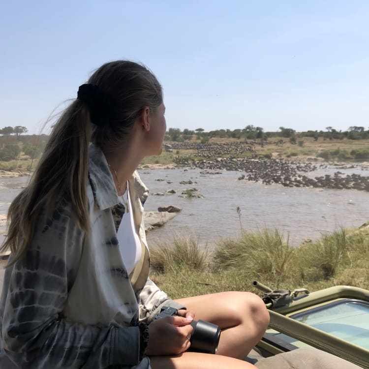 A woman sits near a mobile device, looking out over a river in the Serengeti during the large migration.
