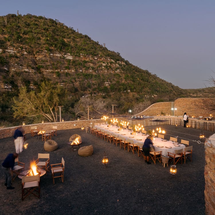 Eine malerische Outdoor-Boma im Abendlicht, die für ein Dinner unter freiem Himmel vorbereitet ist. Lange, weiß gedeckte Tische, umgeben von Stühlen und beleuchtet von Laternen, stehen im Zentrum der Szenerie.