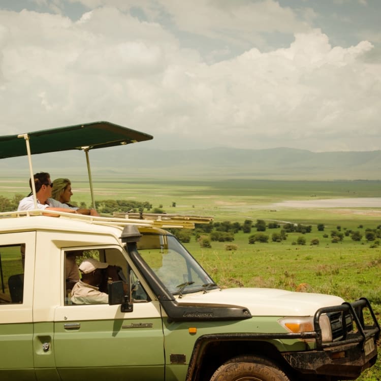 Ein Safarifahrzeug mit Touristen auf einer Safari in einer weiten grasbewachsenen Landschaft in Tansania, mit bewölktem Himmel.