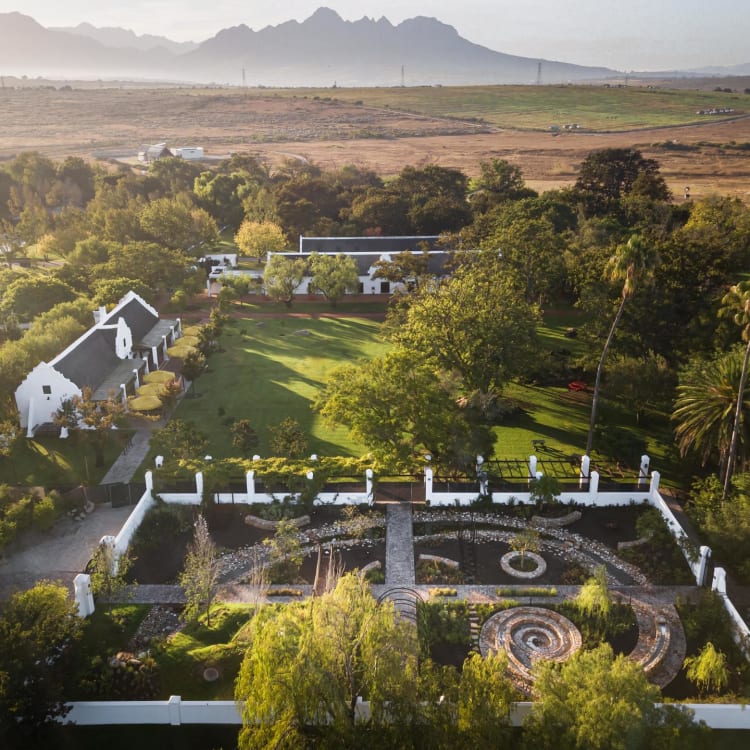 An aerial view of a lush, green estate with white buildings, a central circular garden, and a surrounding white fence, set against a backdrop of mountains.