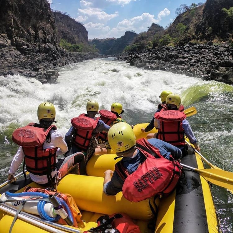 A group of people wearing helmets and life jackets are white water rafting on a turbulent river in a canyon.