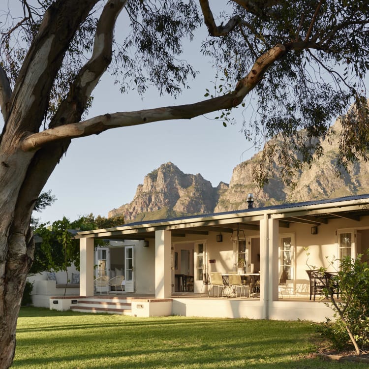 A single-story cottage with a porch, surrounded by green grass and trees, with mountains in the background.