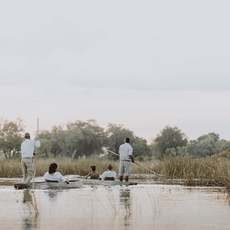 A group of people riding in a traditional mokoro boat through a calm waterway in a lush, green landscape.