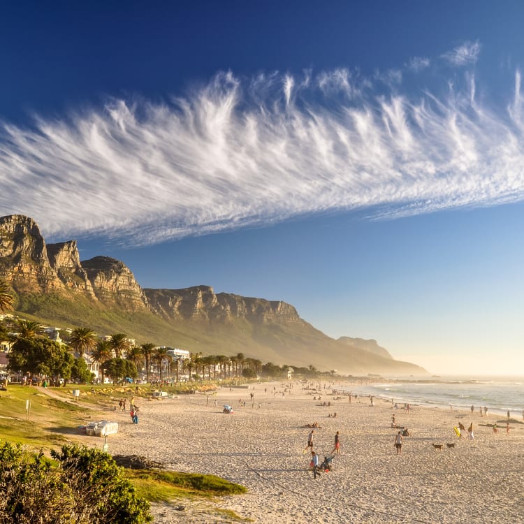 Landschaftlicher Blick auf den Strand von Camps Bay mit Bergen im Hintergrund und Menschen, die am Sand entspannen unter einem teils bewölkten Himmel.