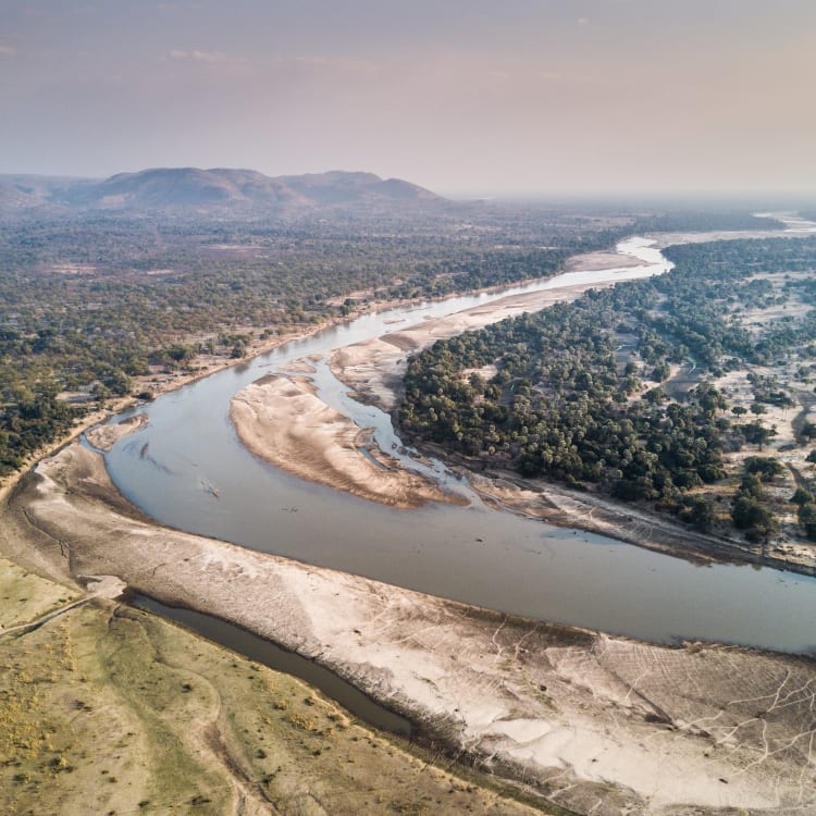 Ein Luftbild des Lower Zambezi Flusses, der durch eine Landschaft mit Wäldern und offene Flächen fließt, mit Hügeln im Hintergrund.