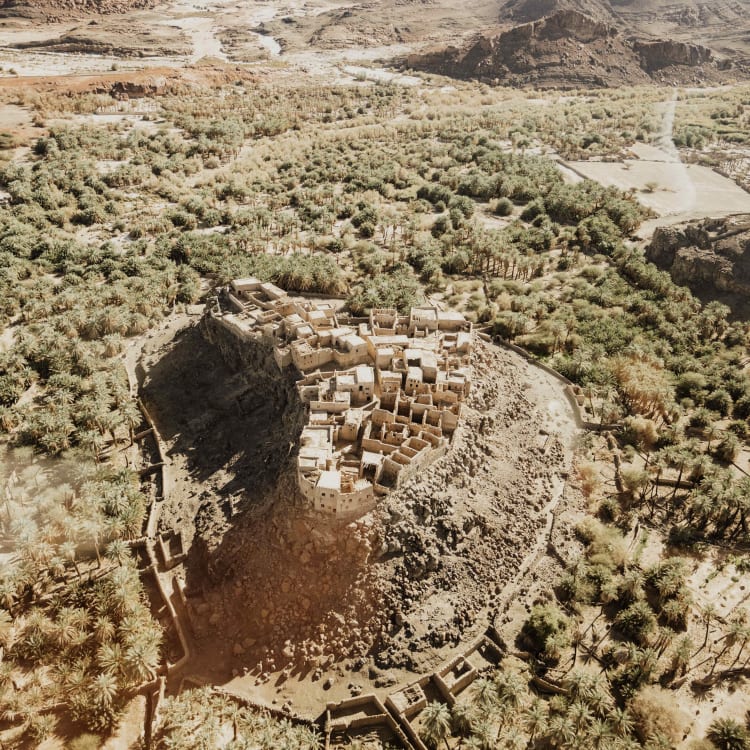 Eine antike Steinfestung, die in einen Hügel gebaut ist und von Grün in einer Wüstenlandschaft umgeben wird.