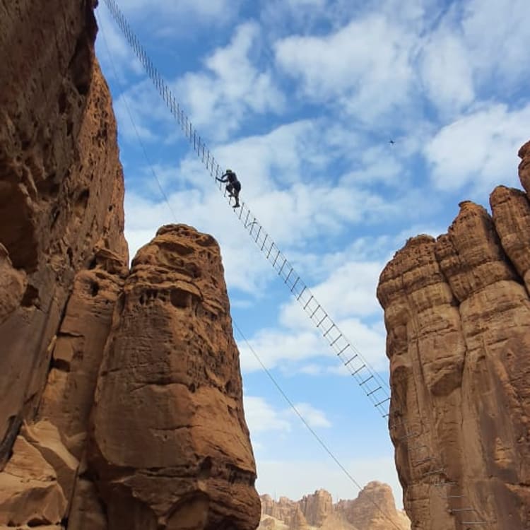 Eine Person steigt auf eine treppenähnliche Brücke zwischen hohen roten Felsen unter einem blauen Himmel mit Wolken.