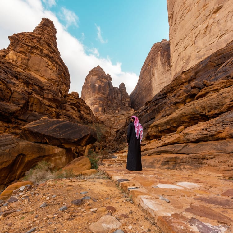 Eine Person steht auf einem felsigen Pfad zwischen steilen Wüstenklippen in Al-Ula, Saudi-Arabien.