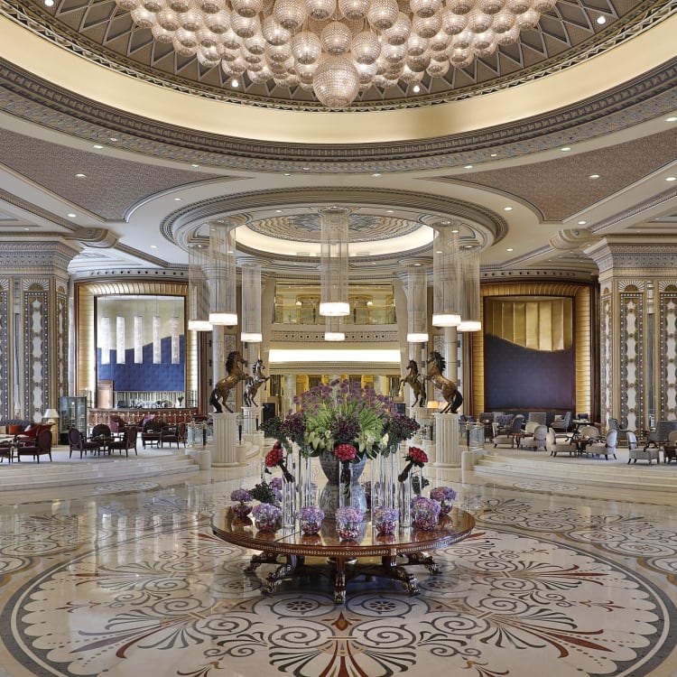 The lobby of a hotel with a marble floor and a table with flowers in the middle under a chandelier