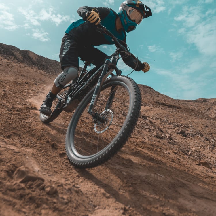 Ein Mountainbiker mit Helm und Schutzkleidung fährt auf einem schmutzigen Weg an einem Hügel unter einem teils bewölkten Himmel.