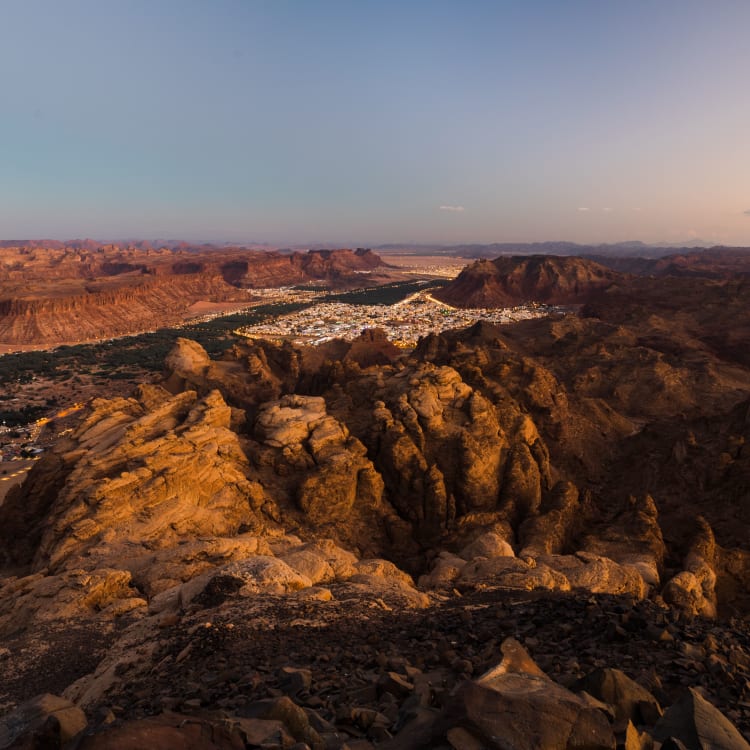 Ein Panoramablick von einem felsigen Aussichtspunkt über eine zerklüftete Wüstenlandschaft mit Bergen und einer kleinen Stadt im Hintergrund bei Sonnenuntergang.