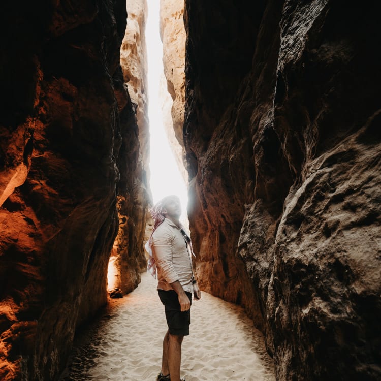 Eine Person steht in einer schmalen, sandigen Schlucht mit hohen Felswänden auf beiden Seiten, die von oben Licht erhalten.