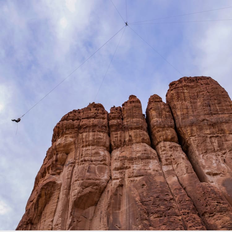 A large rock formation with a giant swing hanging from a line above it, set against a cloudy sky.