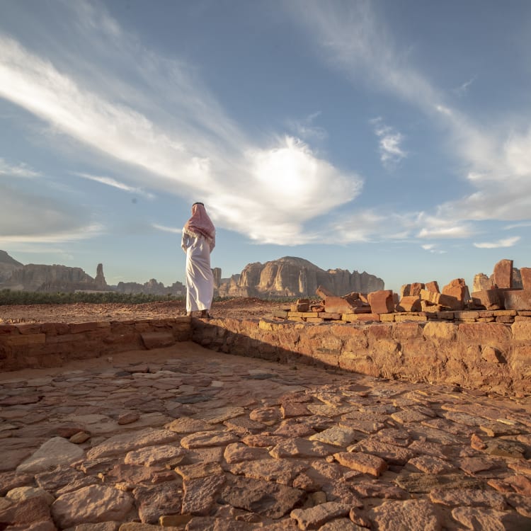 A person dressed in white stands on ancient stone ruins overlooking a desert landscape with distant mountains under a partly cloudy sky.