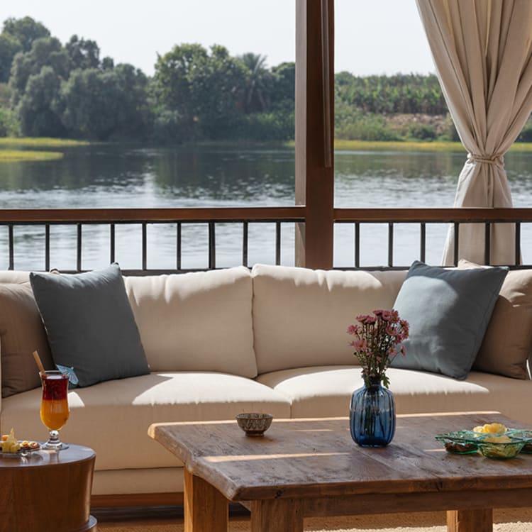 A cozy lounge setup with a white sofa adorned with blue pillows, a wooden coffee table with a blue vase of flowers, and a side table with a drink, overlooking a river and lush greenery under bright daylight.