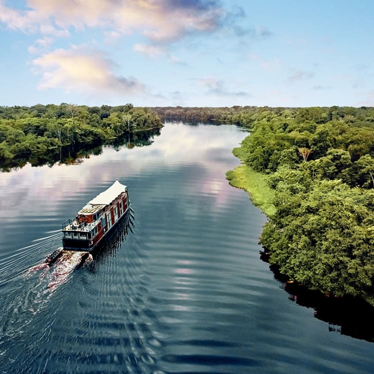 Ein Boot fährt auf einem breiten Fluss durch dicht bewachsenen grünen Regenwald unter einem teils bewölkten Himmel.