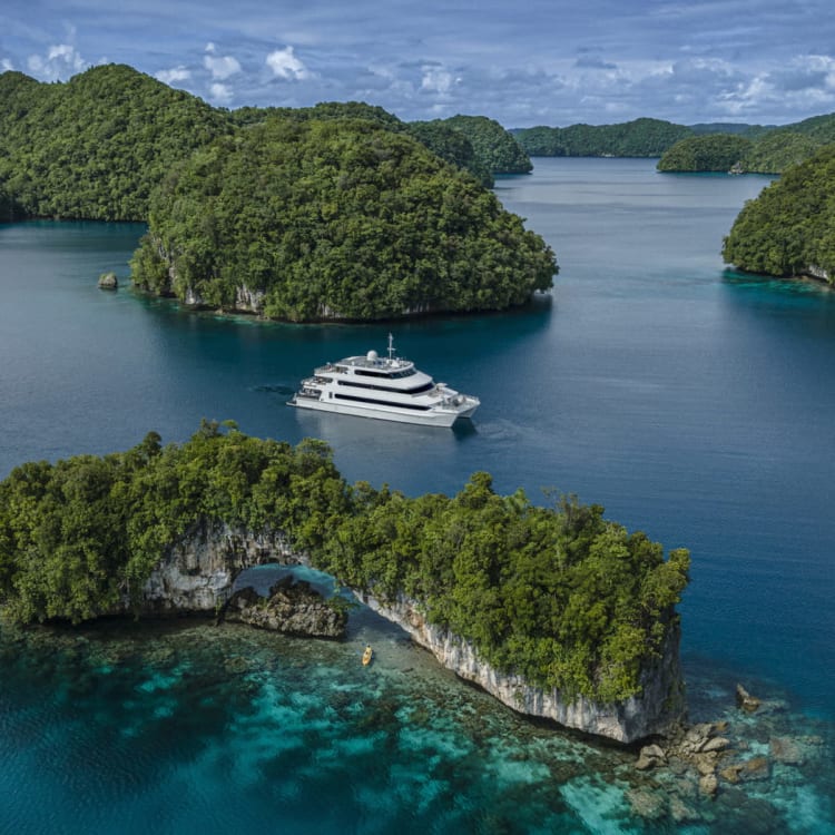 Eine Luxusyacht vor üppig grünen Inseln in einem ruhigen Meer, mit klarem blauem Wasser und partly Wolken am Himmel.