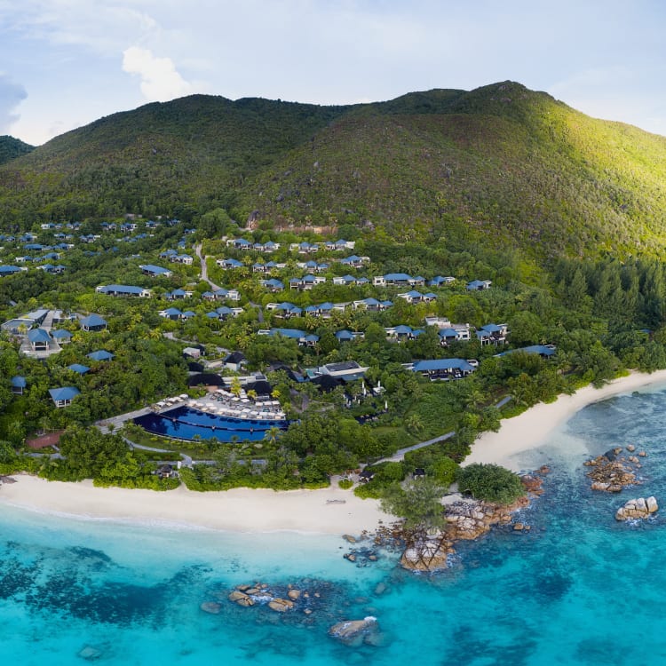 Luftaufnahme des Raffles Seychelles Resorts zwischen grünen Hügeln und türkisfarbenem Ozean, mit weißem Sandstrand und verstreuten Villen mit blauen Dächern.