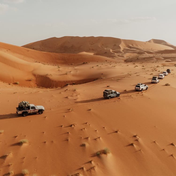 A convoy of cars driving through a vast desert with sand dunes in Oman.