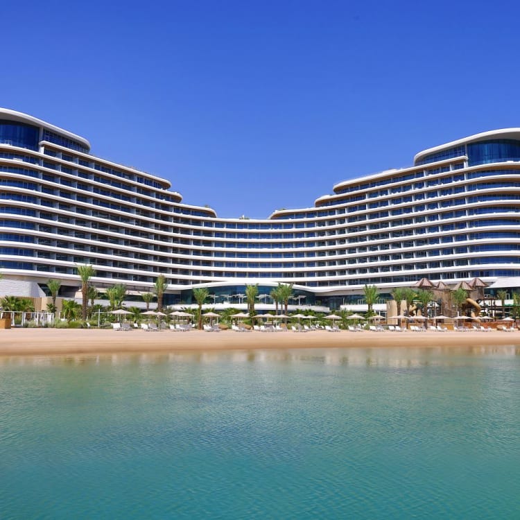 A modern luxury hotel with a curved design, situated by a water body under a clear blue sky.