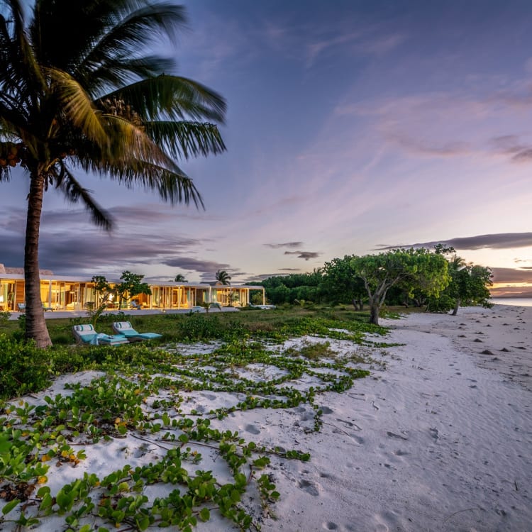Sonnenuntergang an einem tropischen Strand mit Palmen, Sandstrand und einer luxuriösen Villa im Hintergrund.