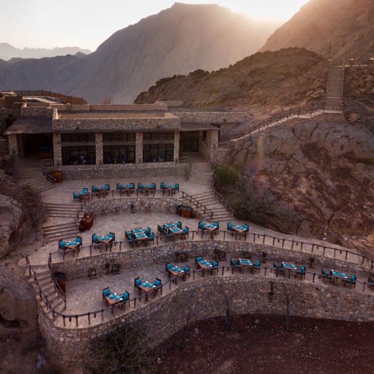 Terrasse mit mehreren Tischen, die auf eine bergige Landschaft in Oman blicken, mit einem modernen Gebäude im Hintergrund.