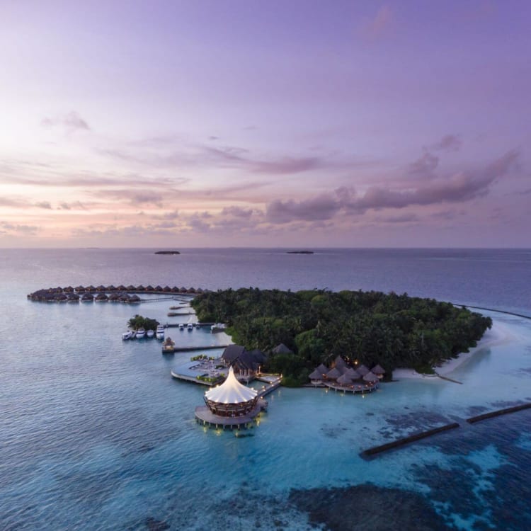 Aerial view of a luxurious island resort in the Maldives during sunset, featuring overwater bavilions, lush greenery, and surrounding turquoise waters.