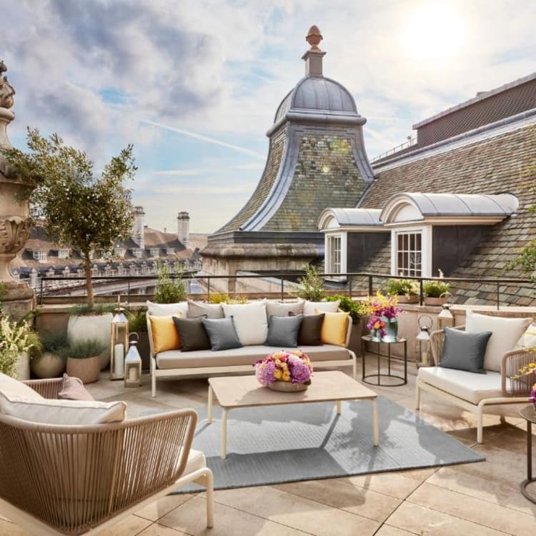 A stylish rooftop lounge with comfortable seating, colorful cushions, and flower arrangements, overlooking historic buildings in London.