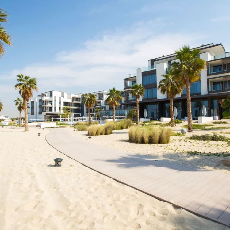 Moderne Wohngebäude am Strand entlang einer Sandpromenade mit Palmen und klarem blauen Himmel.